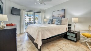 A bedroom with a bed, a TV, a ceiling fan, a nightstand, a chair, and a window with a view of buildings. - CRC 1210 - Second Story Condo with Great Ocean Views (St Augustine)
