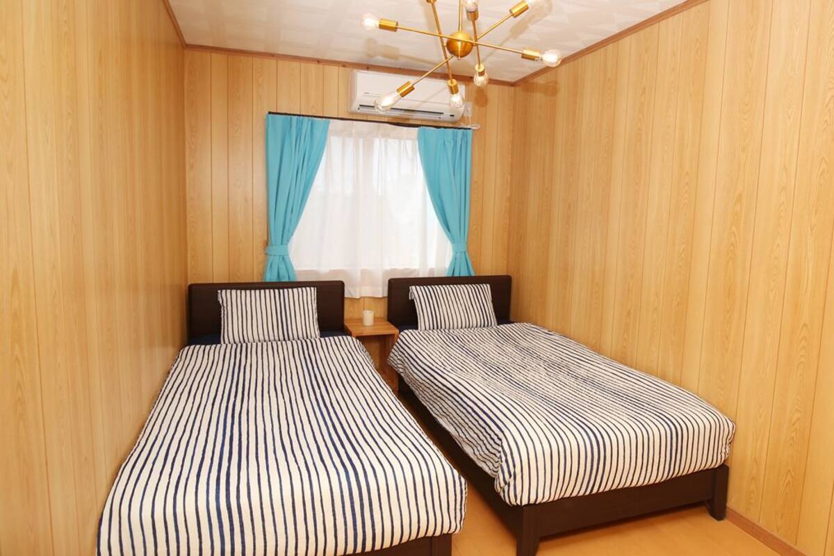 A room with two single beds, wooden walls, a ceiling fan, and a window with curtains.
