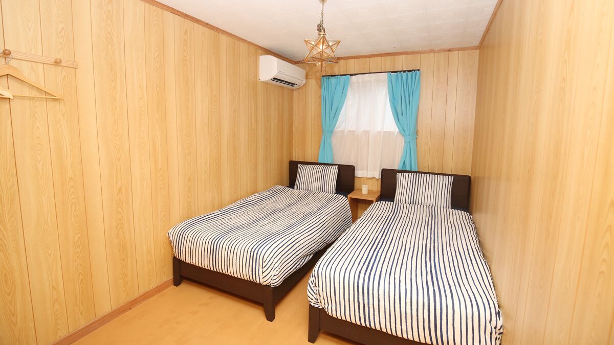 A room with two beds, wooden walls, a hanging light fixture, and a window with curtains.