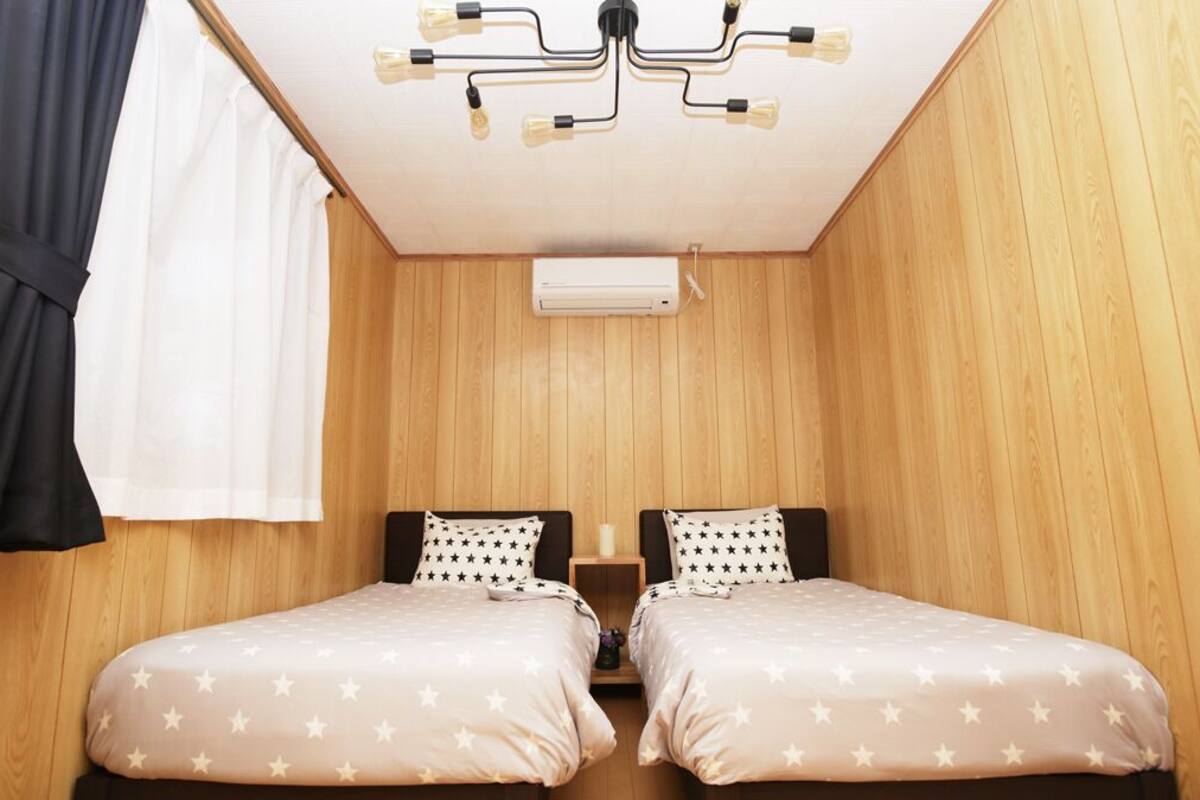 A small, cozy room with wooden walls and a ceiling fan. There are two beds with star-patterned bedding and a small table between them.
