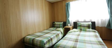 A room with two beds, wooden panel walls, a window with curtains, and a bedside table with a lamp.