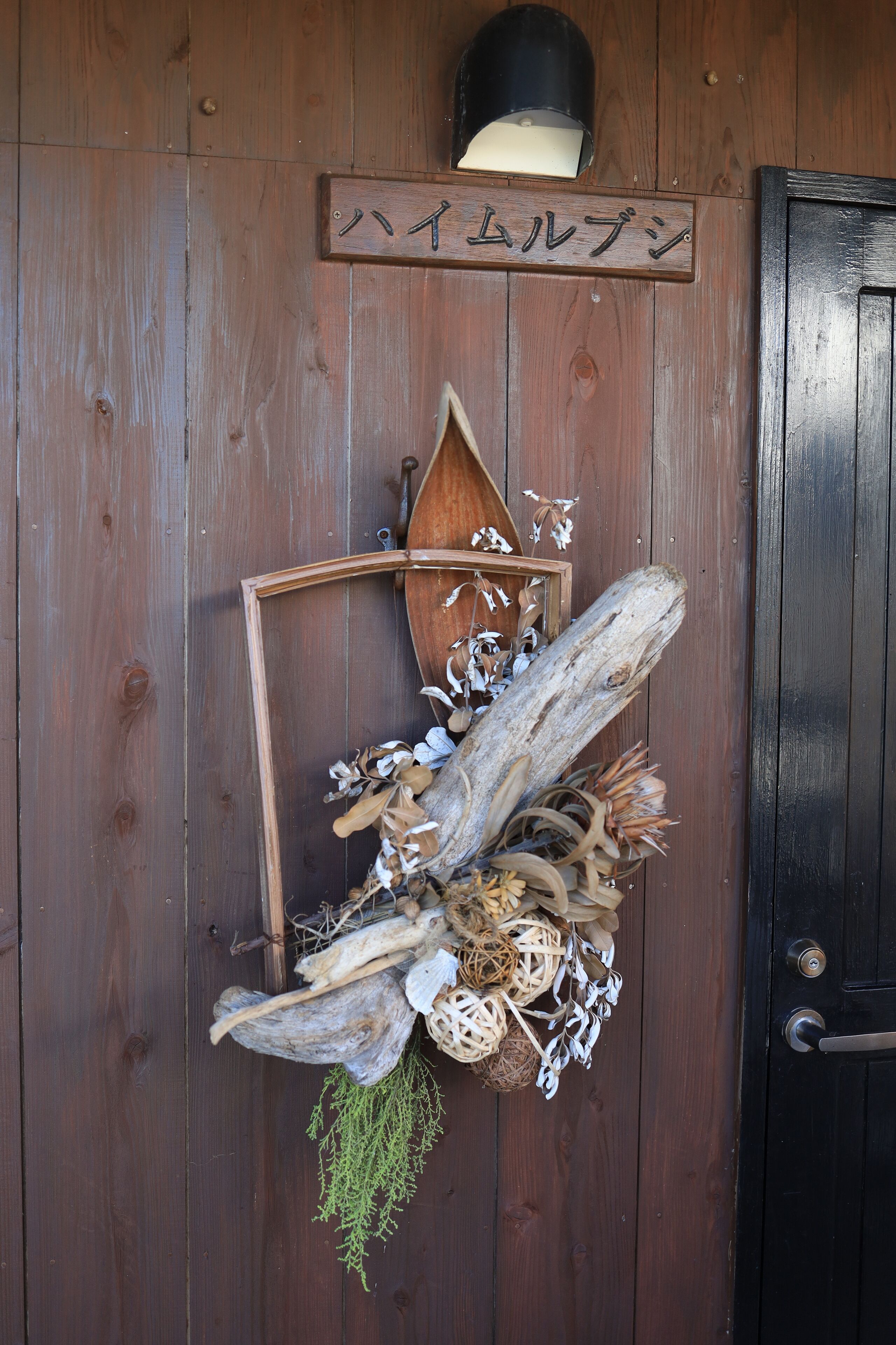 Une porte en bois ornée d’éléments rustiques, notamment une enseigne en bois portant des caractères japonais, une lanterne et divers éléments naturels tels que des feuilles et des branches séchées.