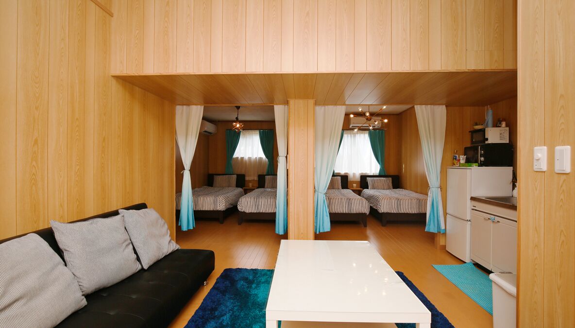 A room with a sofa, a white table, two beds with striped bedding, curtains, a ceiling light fixture, and a small kitchenette.