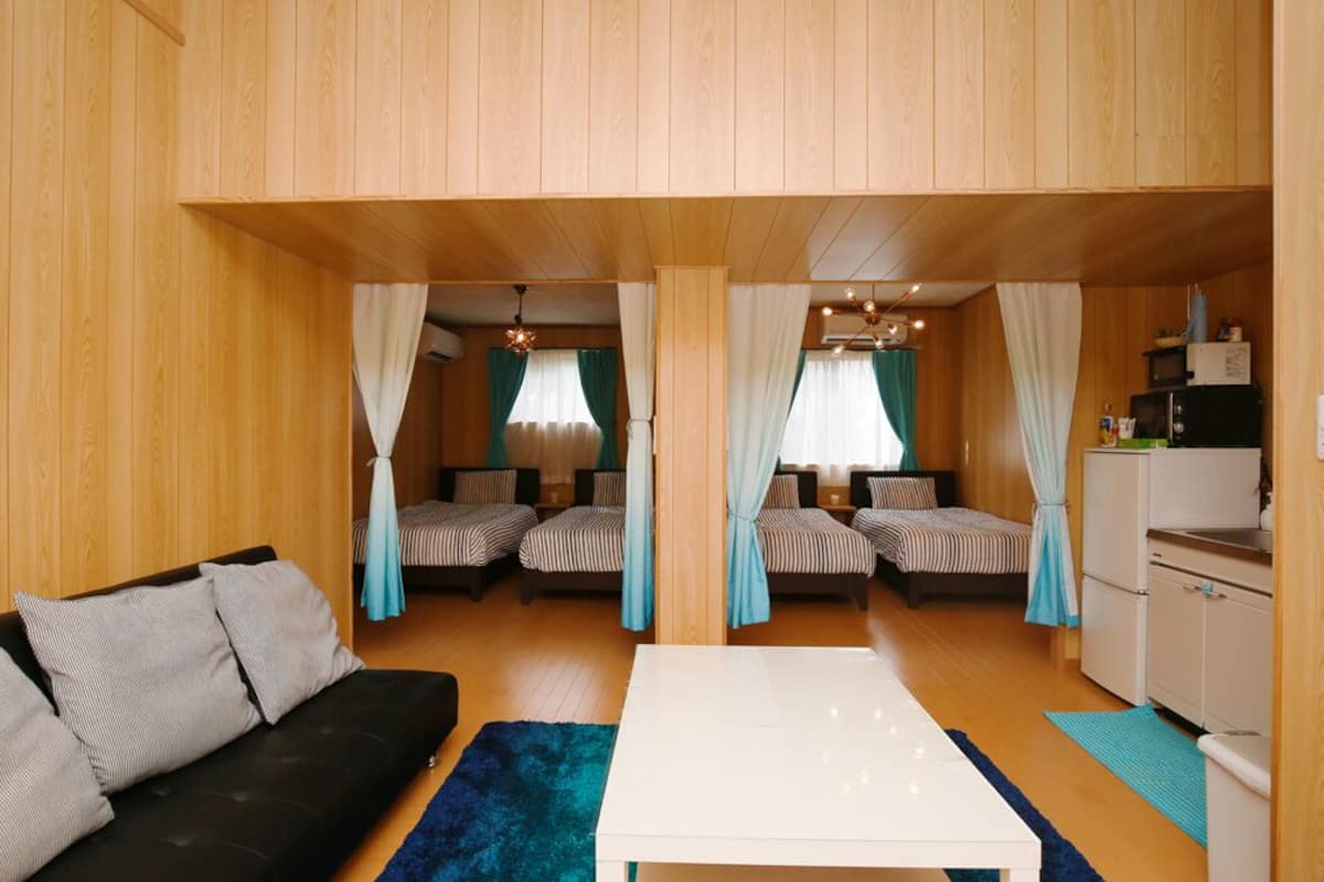 A room with a sofa, a white table, two beds with striped bedding, curtains, a ceiling light fixture, and a small kitchenette.