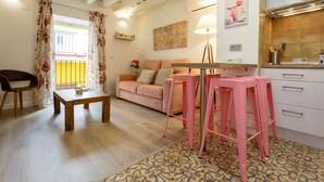 Una sala acogedora con una mesa de madera, un sofá y taburetes rosados. Se ve una zona de cocina con armarios blancos y una alfombra estampada.
