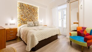 Un dormitorio con una cama, un sillón de colores, una mesita de noche de madera y una pared con un diseño geométrico de azulejos.