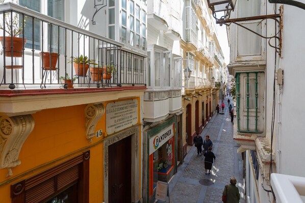 Una calle estrecha flanqueada por edificios con balcones adornados con plantas, una tienda con un letrero y peatones caminando.