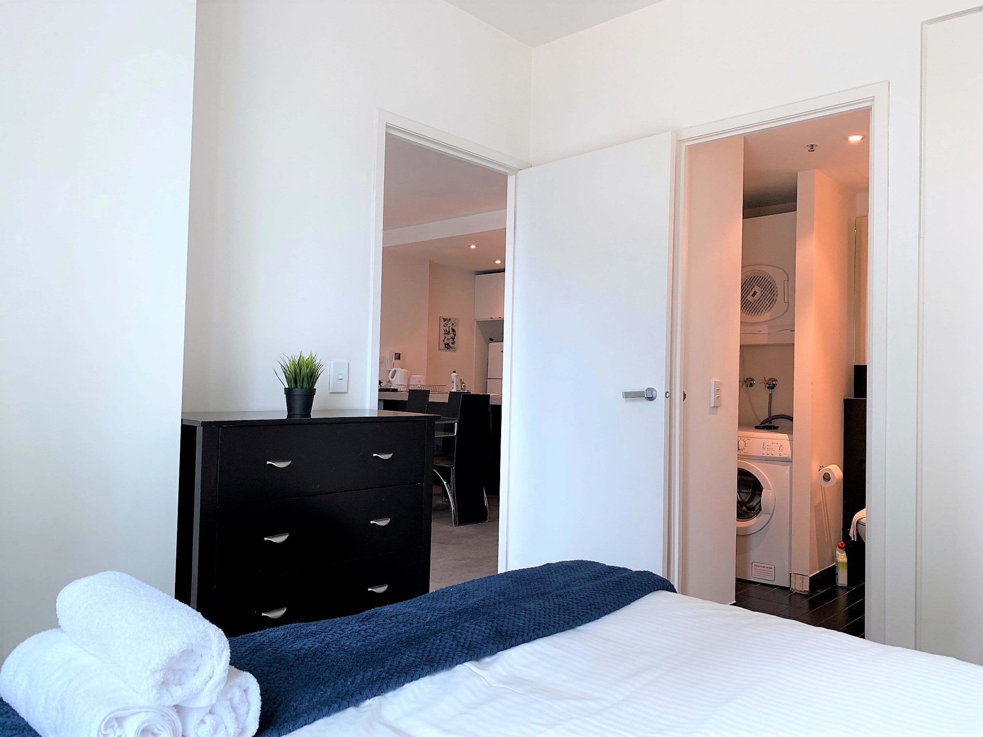 Un dormitorio con cama, cómoda, planta en maceta y vista hacia un cuarto de lavado.
