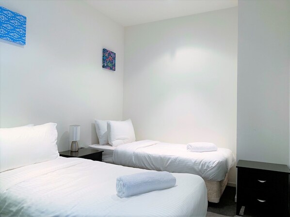 A hotel room with two single beds, white bedding, a small table with a lamp, and a nightstand.
