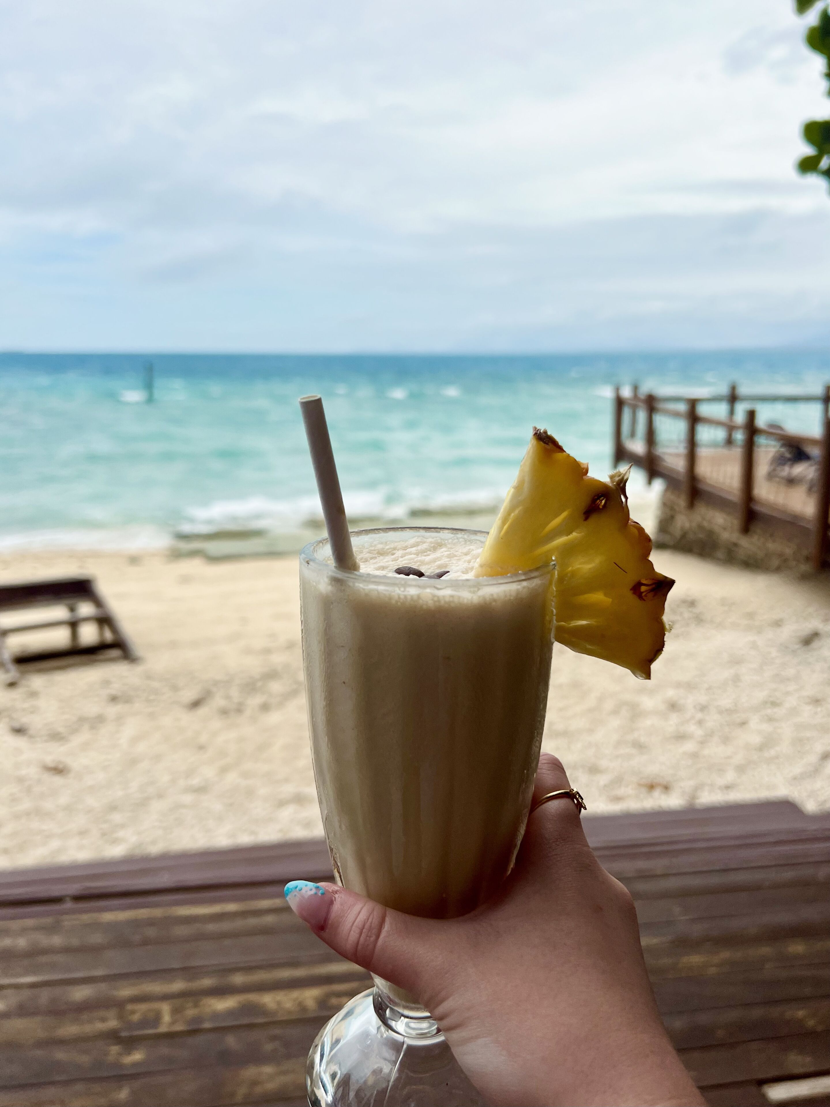 A hand holding a glass with a straw and a pineapple slice, overlooking a beach with a wooden fence and a bench.