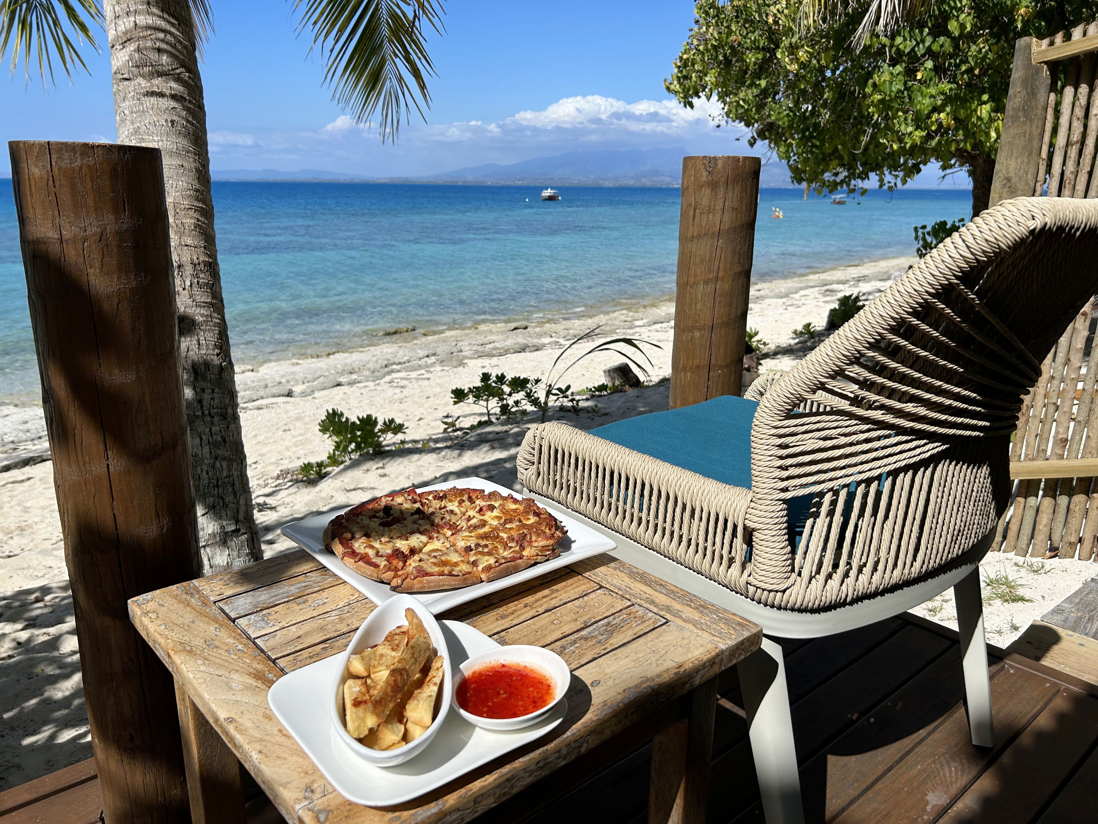 A wooden table with a pizza, a bowl of chips, and a bowl of sauce, set on a wooden deck overlooking a beach with a clear view of the ocean and a boat in the distance.