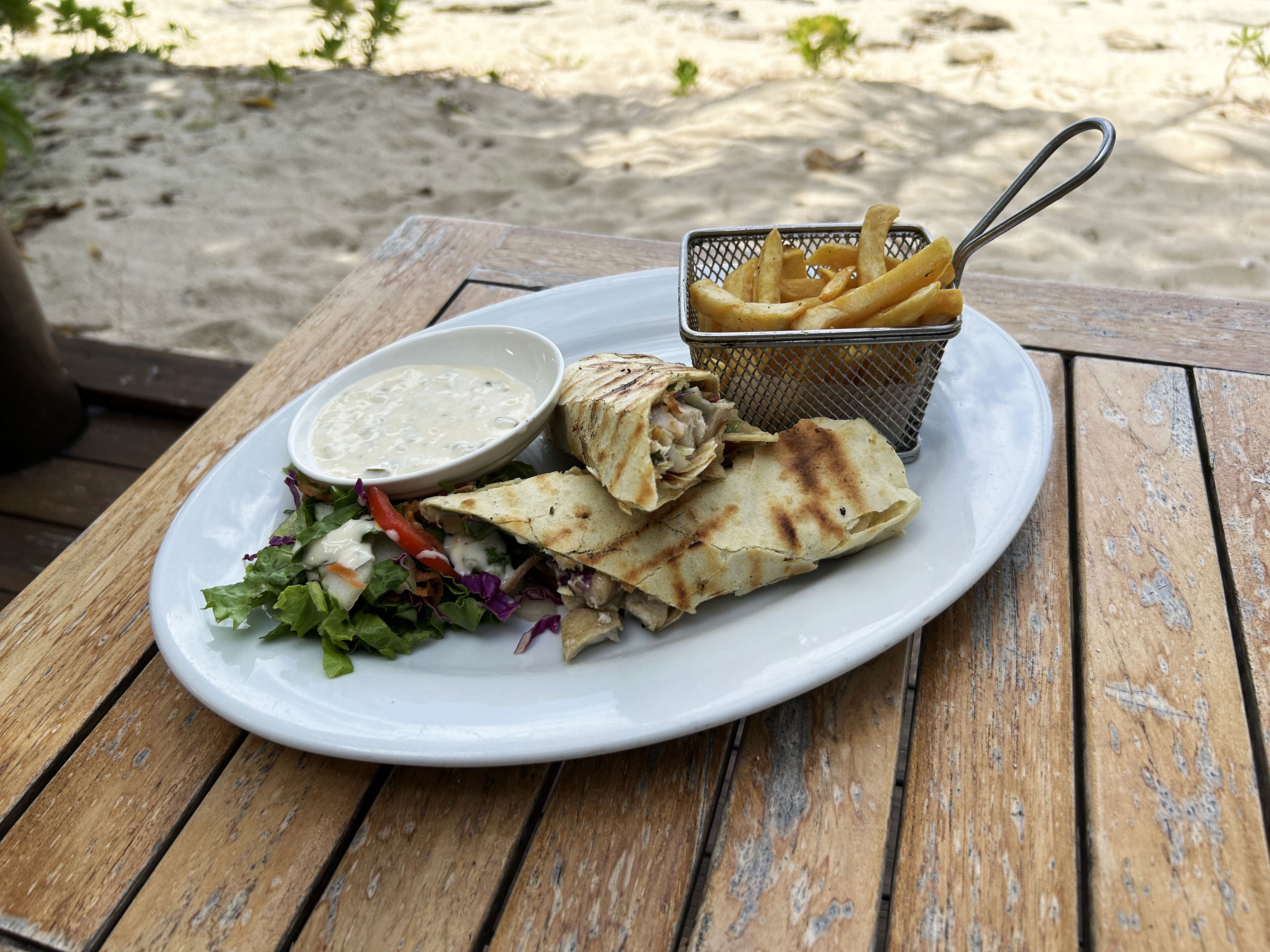 A plate with a wrap, fries, and a salad with a dipping sauce on a wooden table.