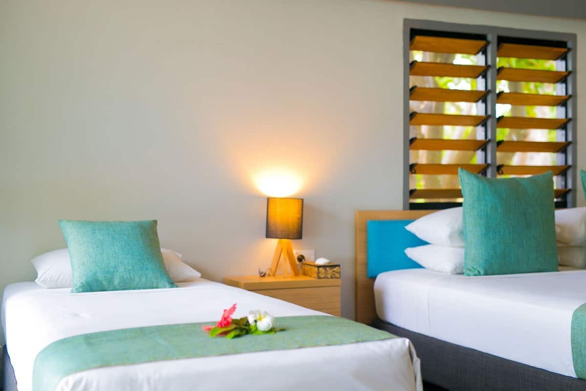 A hotel room with two beds, a nightstand with a lamp, and a window with blinds.