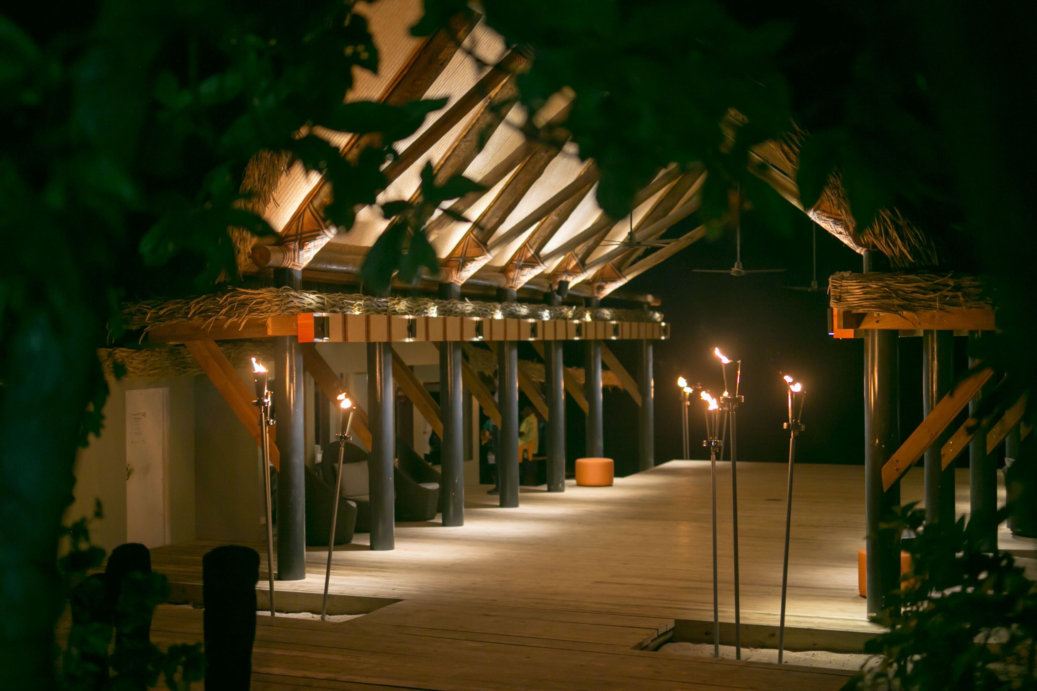 A covered outdoor area with wooden beams and pillars, a thatched roof structure, and several torches providing light.
