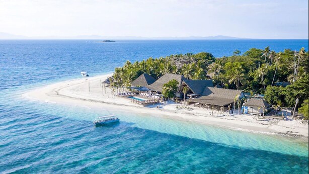 An aerial view of a beach resort with clear turquoise waters, a sandy beach, and thatched-roof buildings.