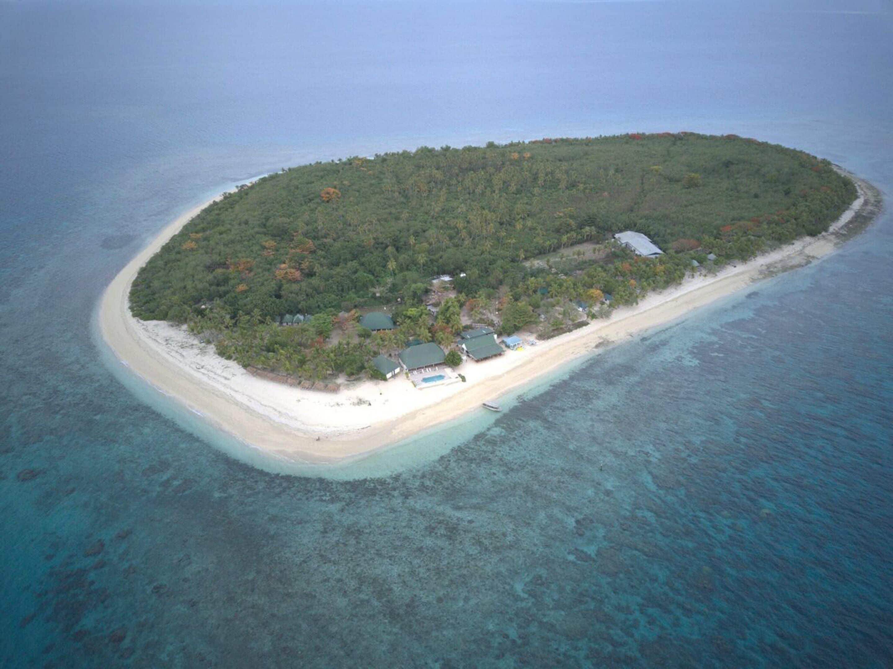 An aerial view of a small island with a sandy beach, dense forest, and several buildings with roofs.