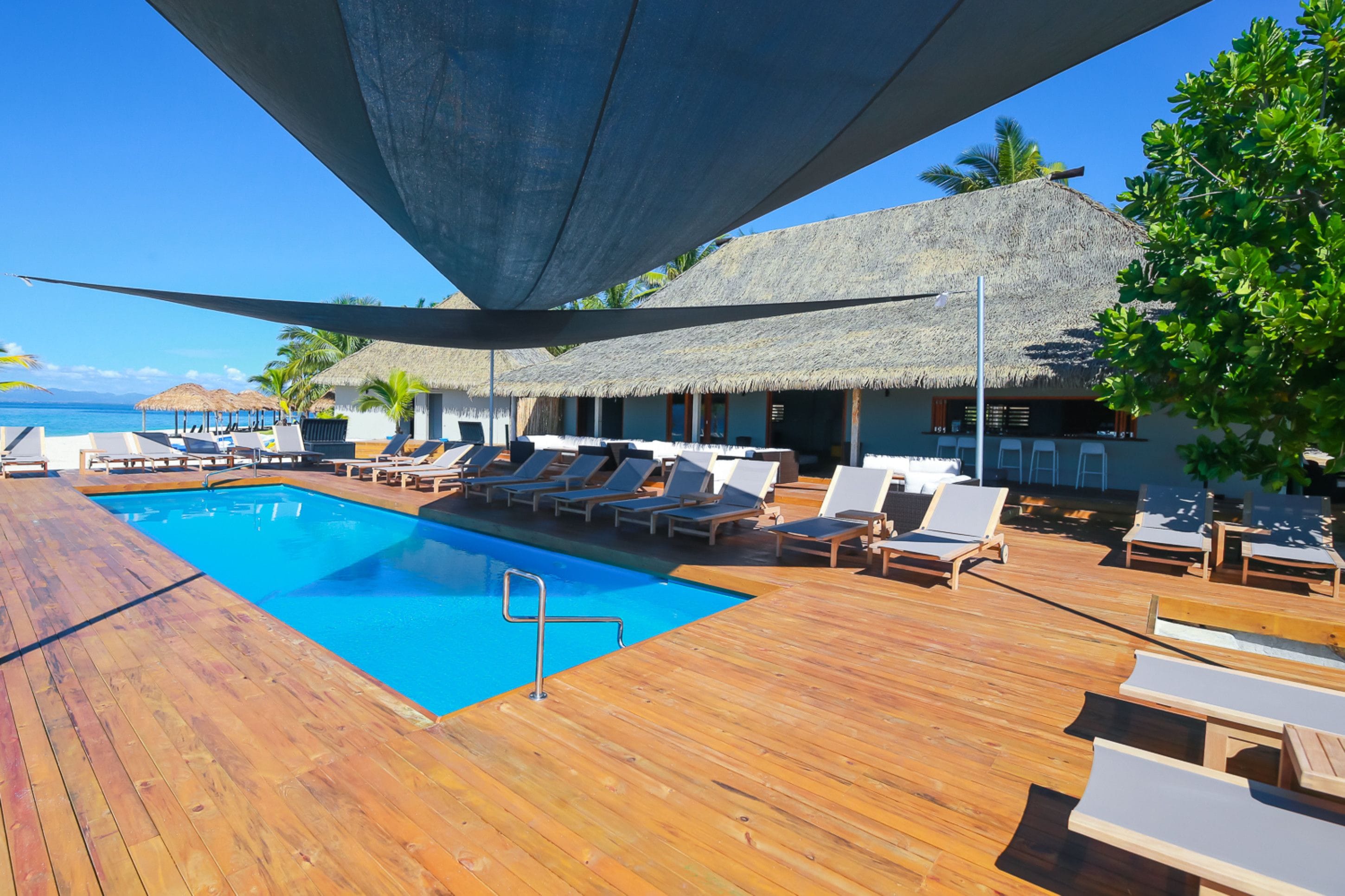 Una terraza de madera con piscina, sillas de descanso y edificios con techo de paja.
