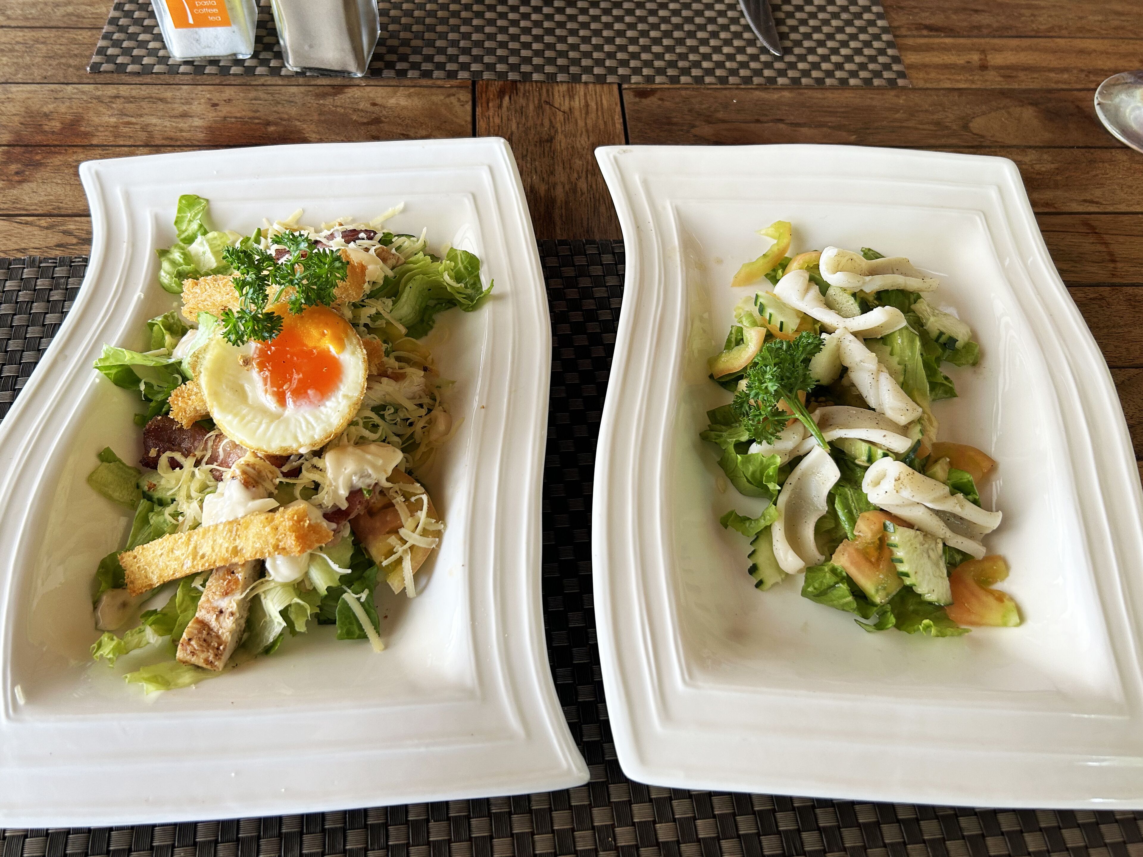 Dos platos rectangulares blancos con ensalada sobre una mesa de madera.