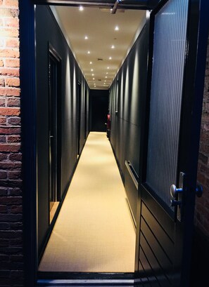 Un couloir étroit, dont le sol est recouvert d’une moquette, bordé de murs et de portes sombres, et qui mène à une pièce plongée dans la pénombre au fond.