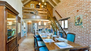 A dining area with a long wooden table set for six, flanked by blue upholstered chairs. A brick wall and wooden staircase are visible in the background.