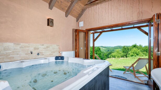 A hot tub with a stone-tiled wall, a wooden ceiling, and a view of a green landscape through an open door.
