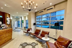 A hotel lobby with a reception desk, leather seating, a chandelier, and large windows with a view of buildings.