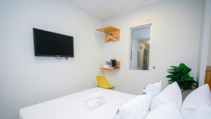 Une chambre d’hôtel avec un lit, un téléviseur fixé au mur, une petite étagère en bois avec des articles de toilette, un miroir et une plante en pot.