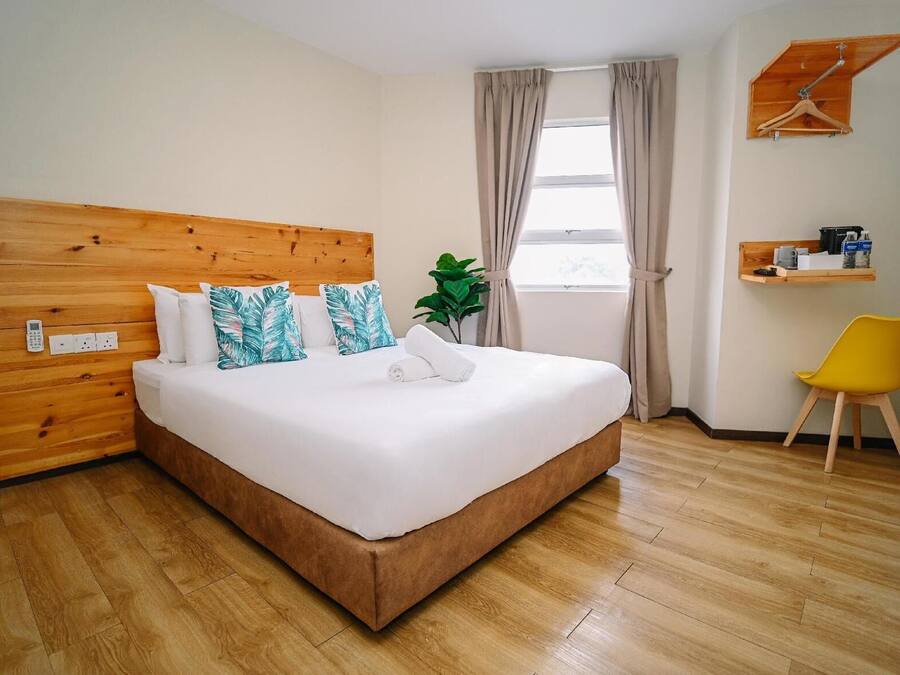 A hotel room with a wooden headboard, a bed with white bedding, a plant, a window with curtains, a wooden shelf with items, and a yellow chair.