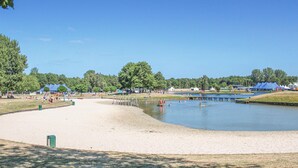 Ein Sandstrandabschnitt mit einem ruhigen See, umgeben von Bäumen und Zelten, vermutlich ein Erholungsgebiet.