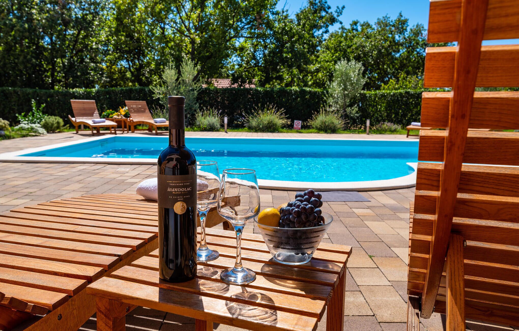 A wooden table with a bottle of wine, a glass, and a bowl of grapes by a pool.