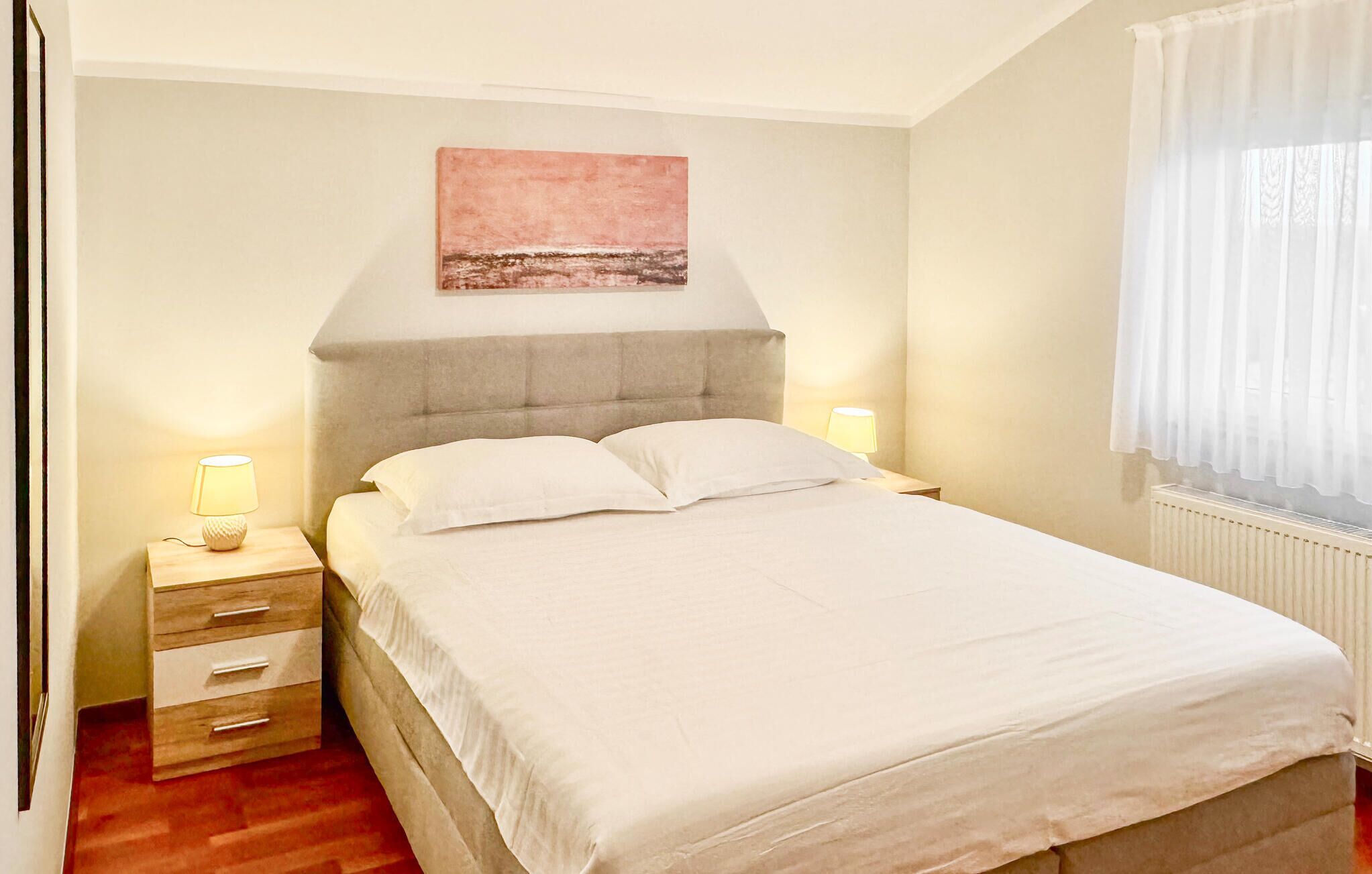 A bedroom with a bed, bedside lamps, a nightstand, a wall-mounted painting, and a window with curtains.