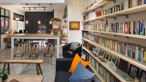 Un rincón acogedor para leer, con un sofá negro, taburetes de madera y una estantería llena de libros.