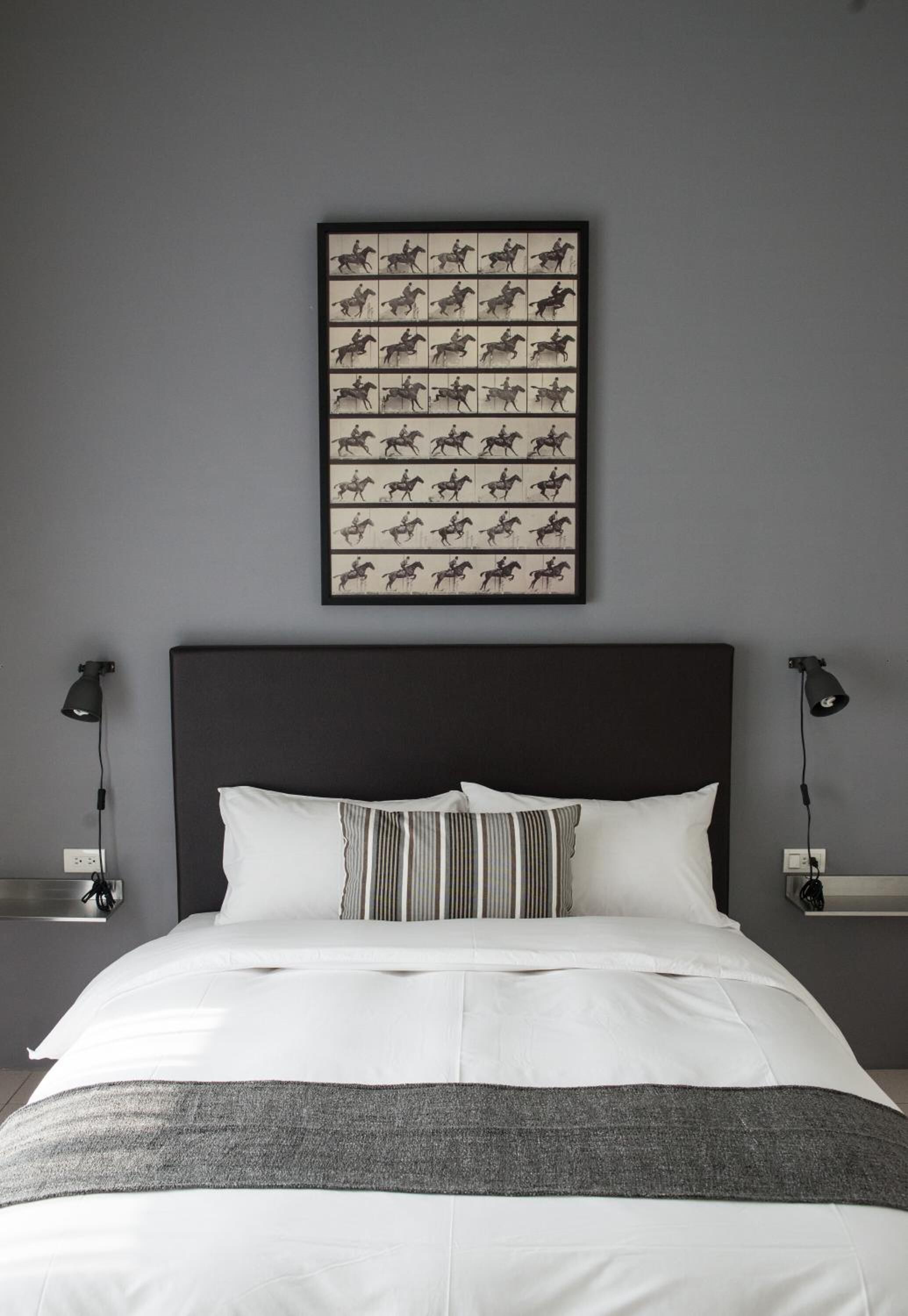 A neatly made bed with white linens and a gray throw blanket, a framed picture of horses on the wall, and two bedside lamps.