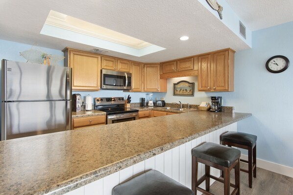 A kitchen with granite countertops, wooden cabinets, a stainless steel refrigerator, a microwave, and a clock on the wall.