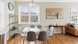 Un comedor con una mesa de vidrio, sillas grises, un soporte metálico para planta y un cuadro de un árbol.