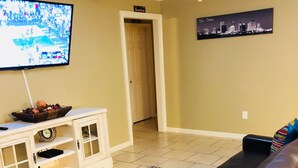 A living room with a TV, a white cabinet, a black leather couch, and a cityscape painting.