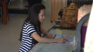 A person sitting at a table with a notebook and pen, in a room with a wooden statue and a chair in the background. - Blue Clouds Homestay (Da Nang)