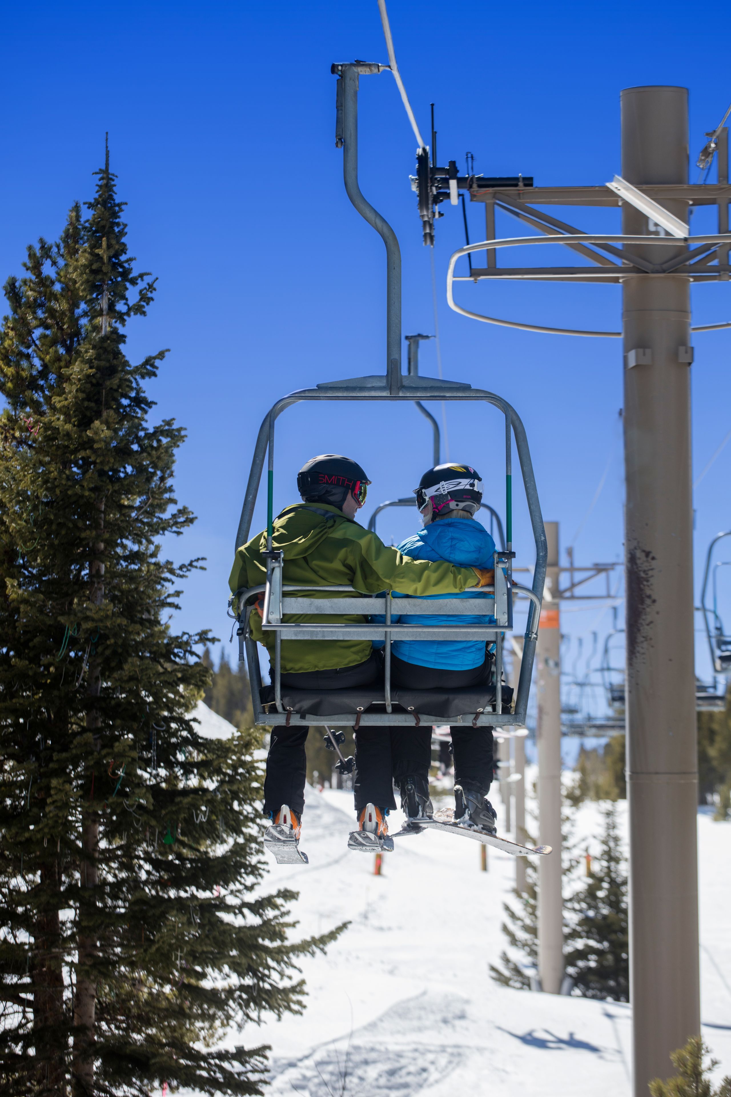 Deux personnes dans un télésiège, entourées de neige et d’arbres.