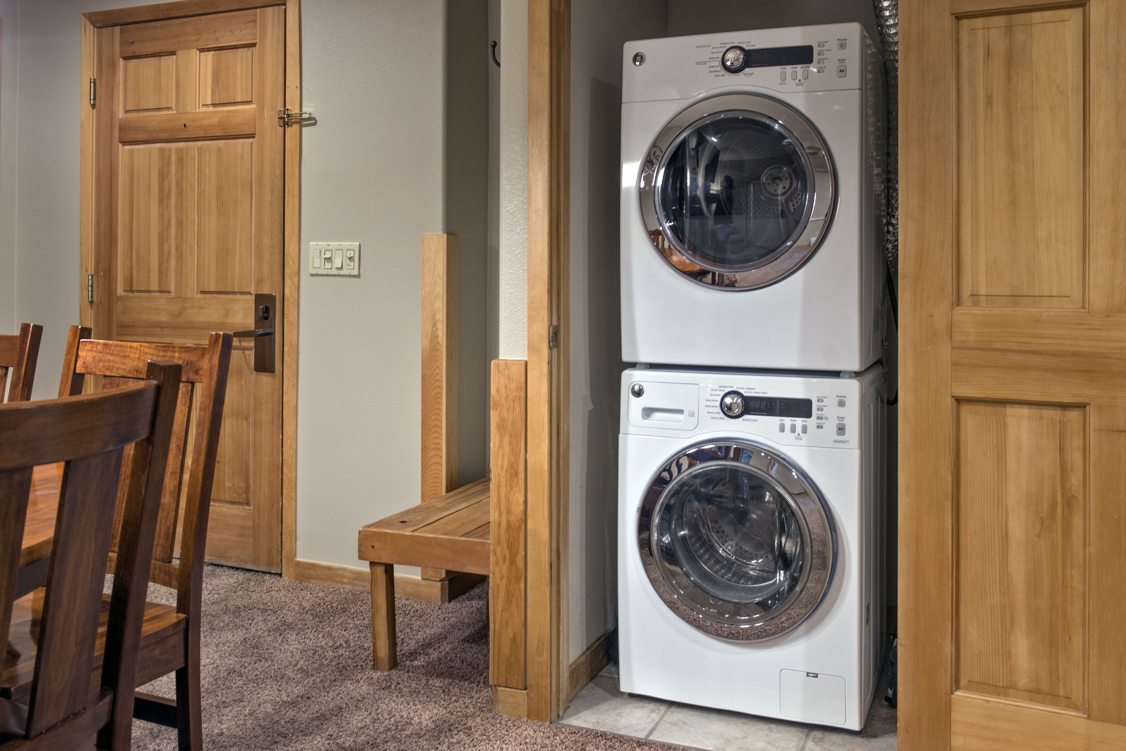 A laundry room with two stacked front-loading washing machines, wooden doors, a small wooden bench, and a carpeted floor.