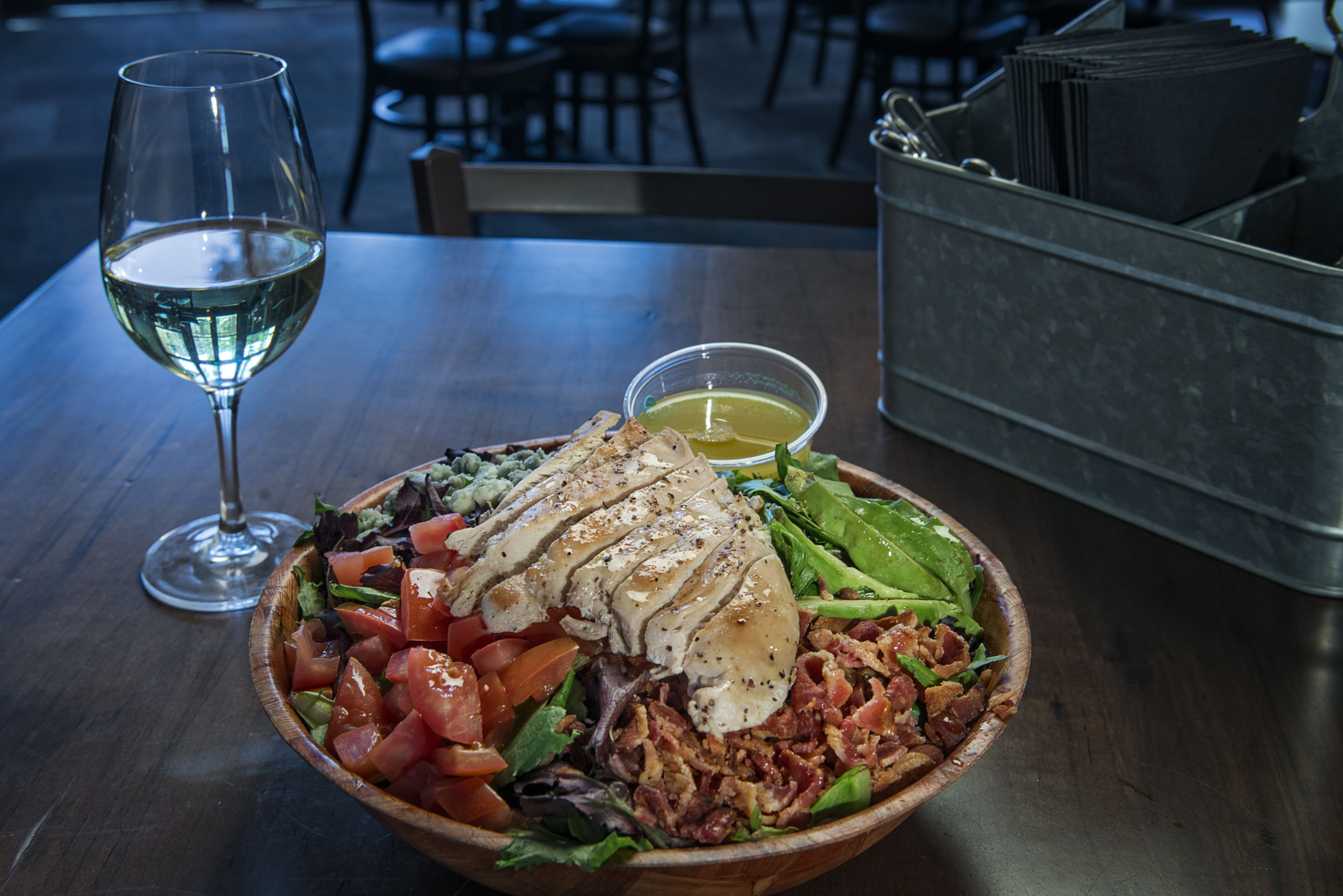 A wooden bowl of salad with a glass of white wine on a table.