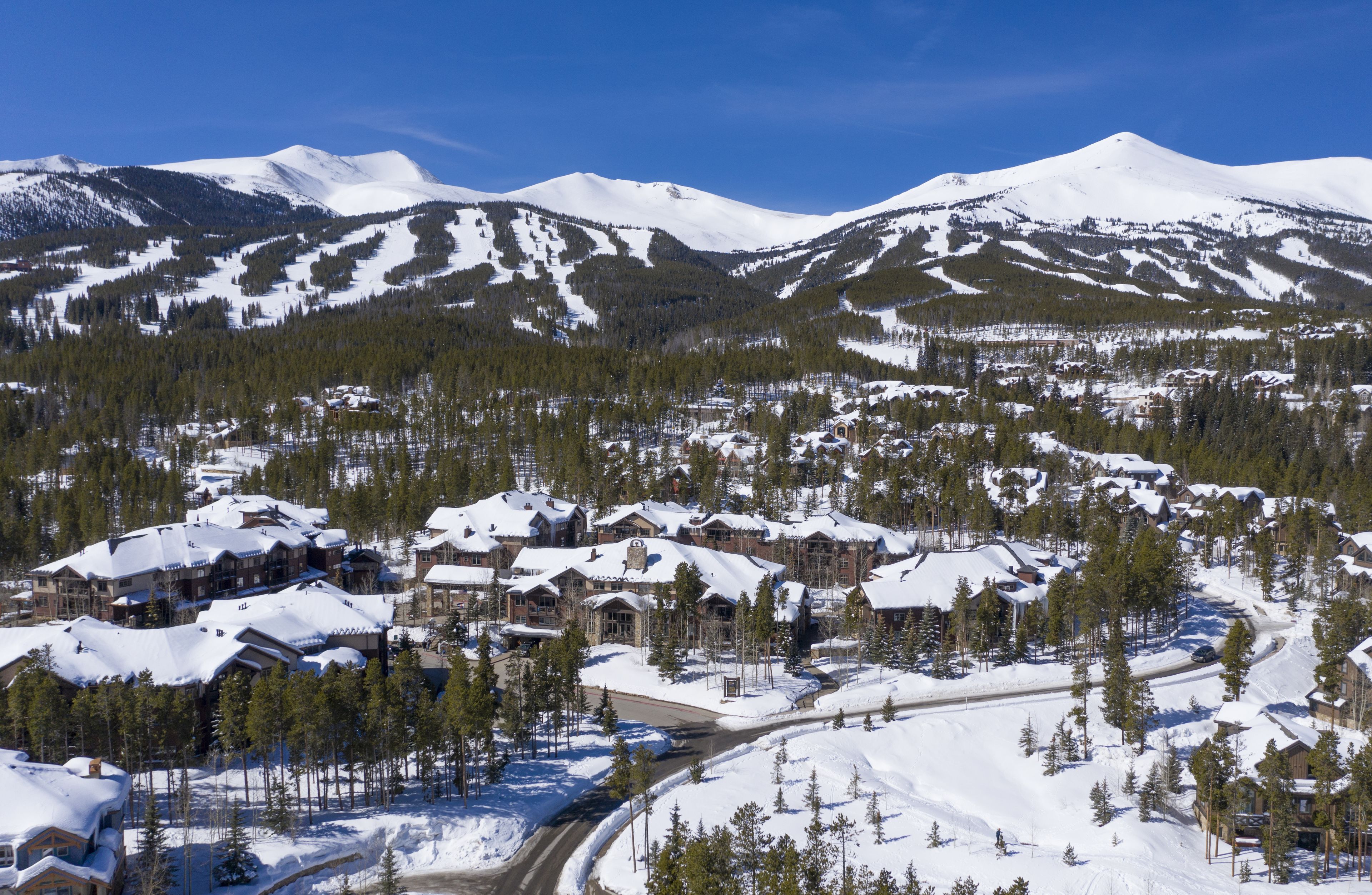 Une station de montagne enneigée, dotée de plusieurs bâtiments d’hébergement, de pistes de ski et d’un ciel d’un bleu éclatant.