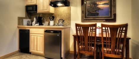 A compact kitchen with wooden cabinets, a built-in microwave, a coffee maker, and a refrigerator. There is a dining table with chairs and a framed picture on the wall.