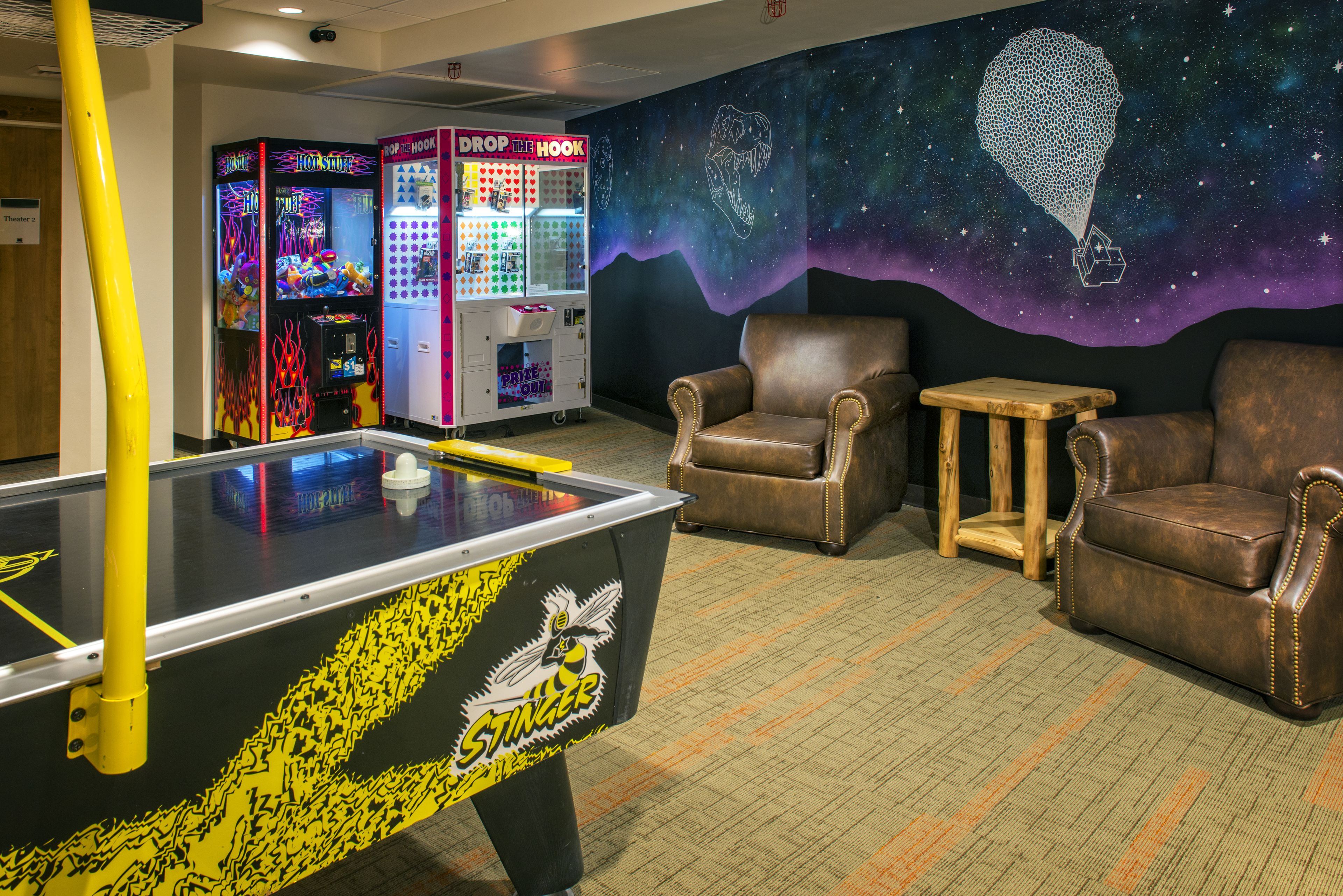 A pool table with a yellow and black design, two claw machines, and a mural of a hot air balloon in a room with leather chairs and a small table.