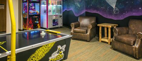 A pool table with a yellow and black design, two claw machines, and a mural of a hot air balloon in a room with leather chairs and a small table.
