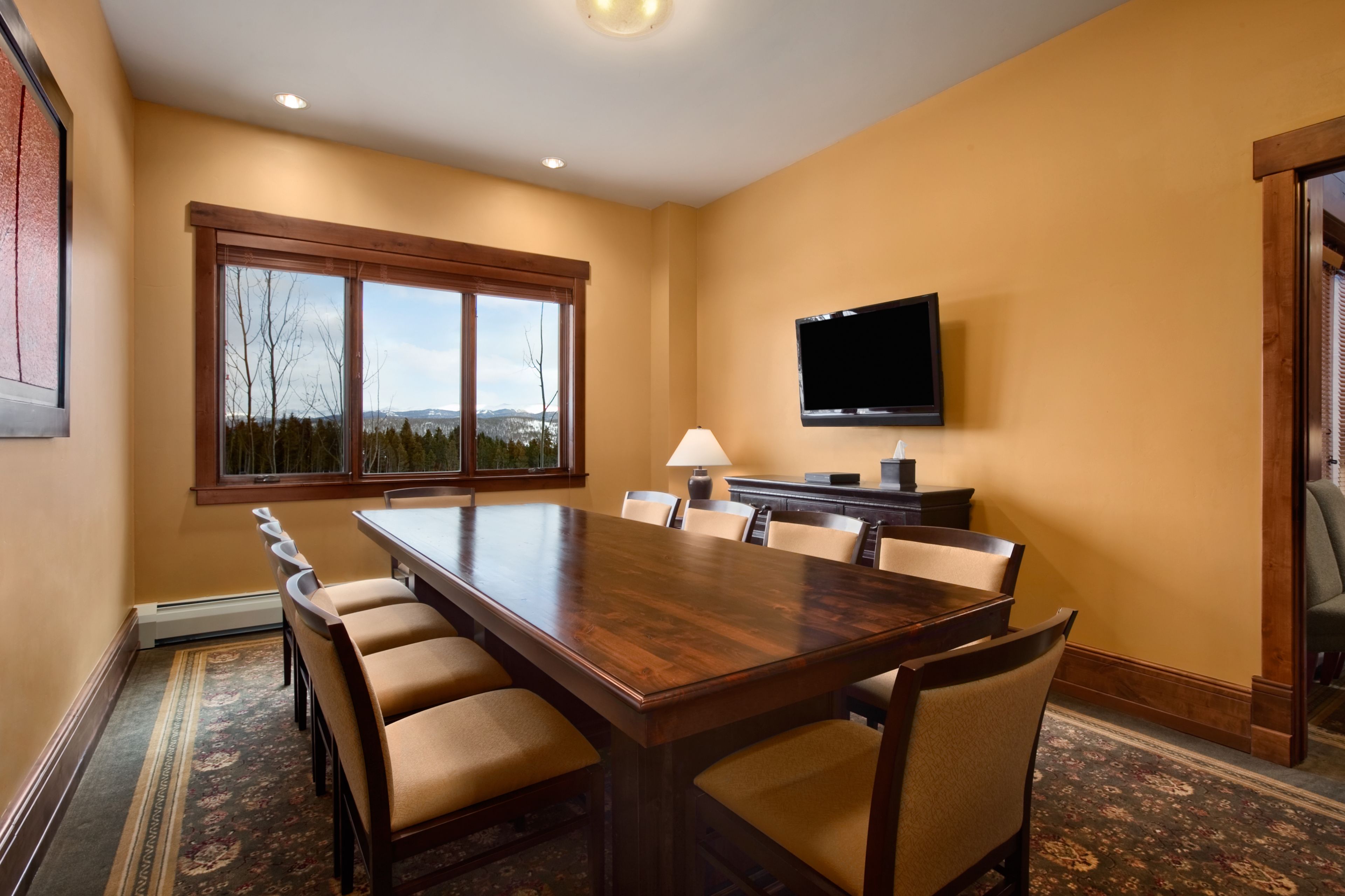 A conference room with a large wooden table, beige chairs, a television mounted on the wall, and a large window with a view of trees and mountains.
