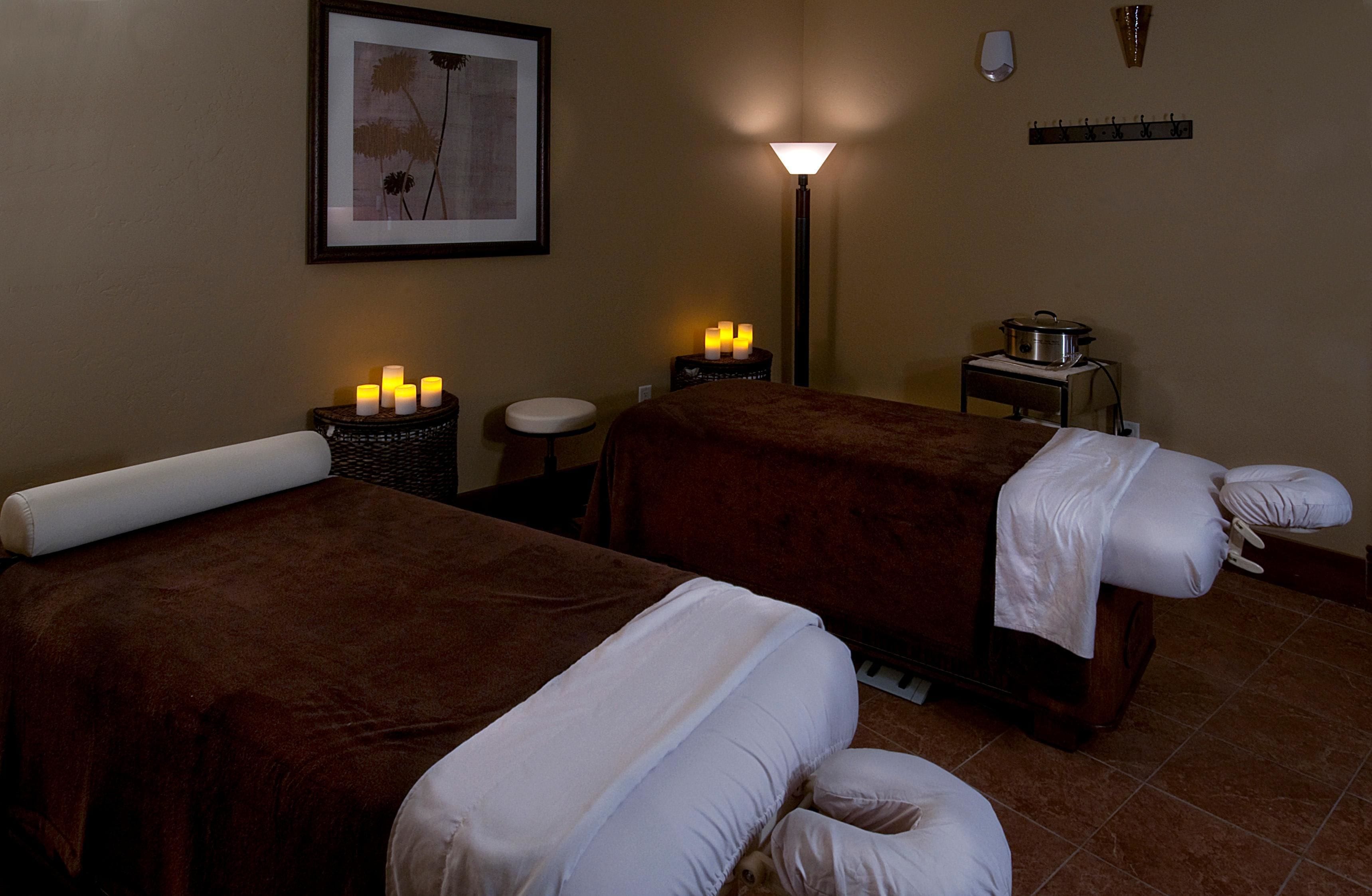 A room with two massage beds, a small table with candles, a floor lamp, and a wall-mounted shelf.