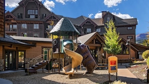 Un terrain de jeux avec un toboggan et des escaliers, un bâtiment en bois doté d’une façade en pierre, et un ciel bleu dégagé.