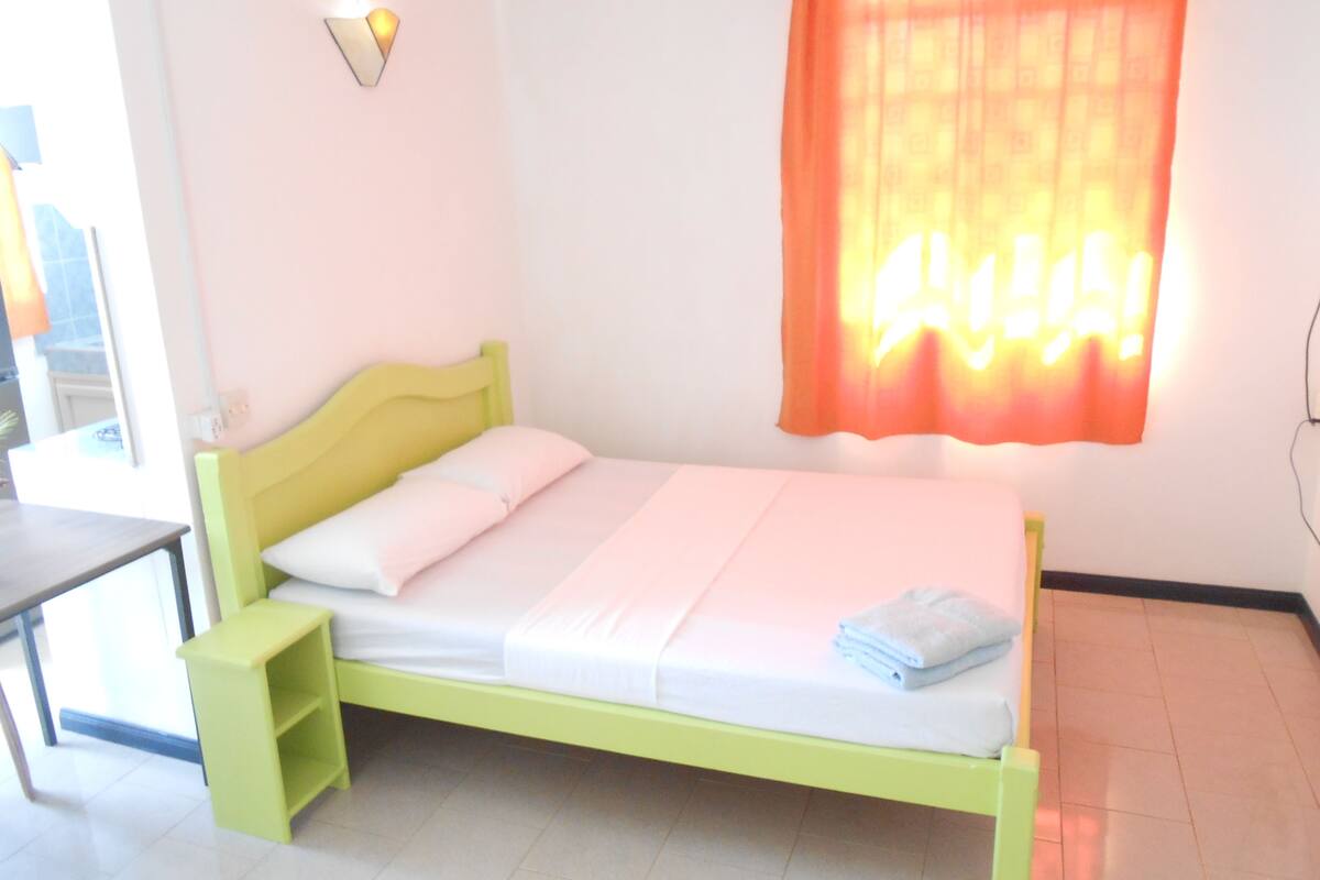 Una cama con estructura verde y ropa de cama blanca, una mesita de noche y una lámpara fijada a la pared.