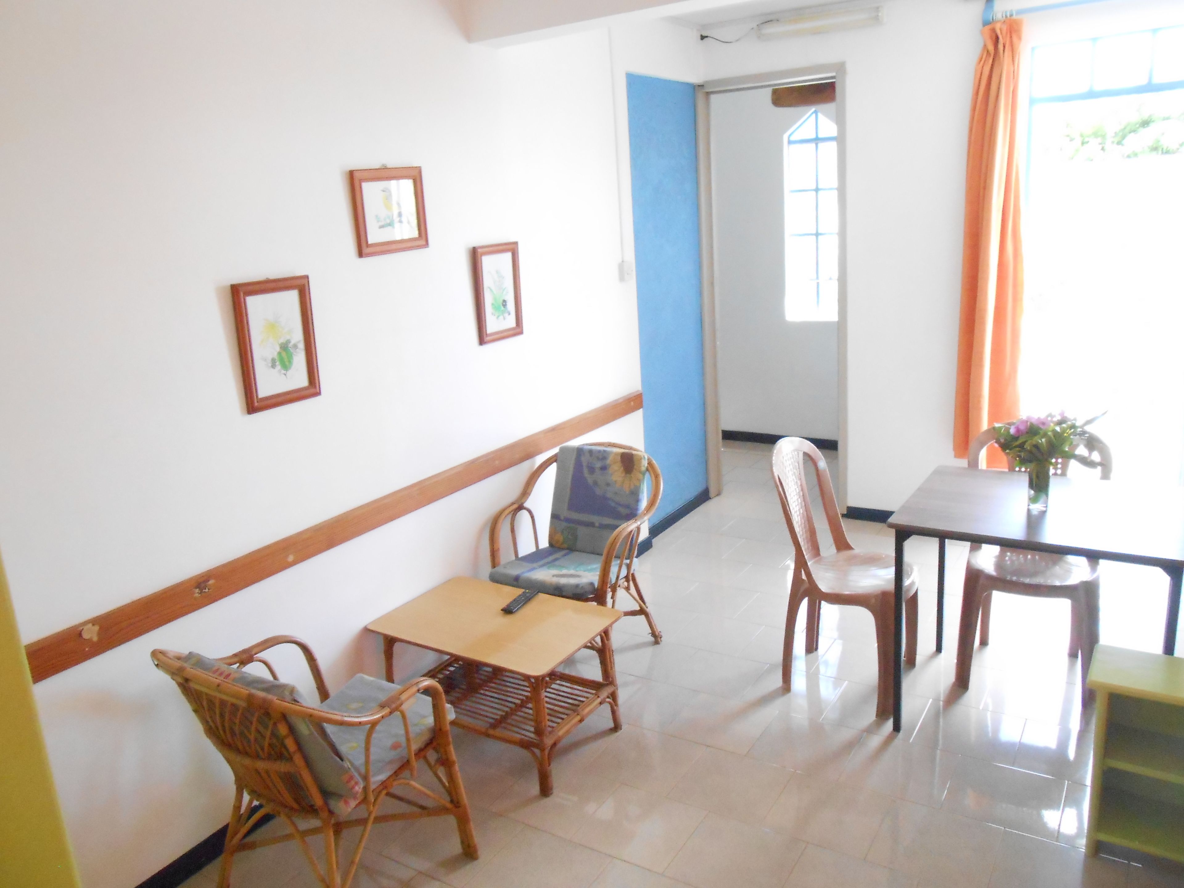 A room with a dining table, chairs, and a small table with a vase. There are framed pictures on the wall and a window with curtains.