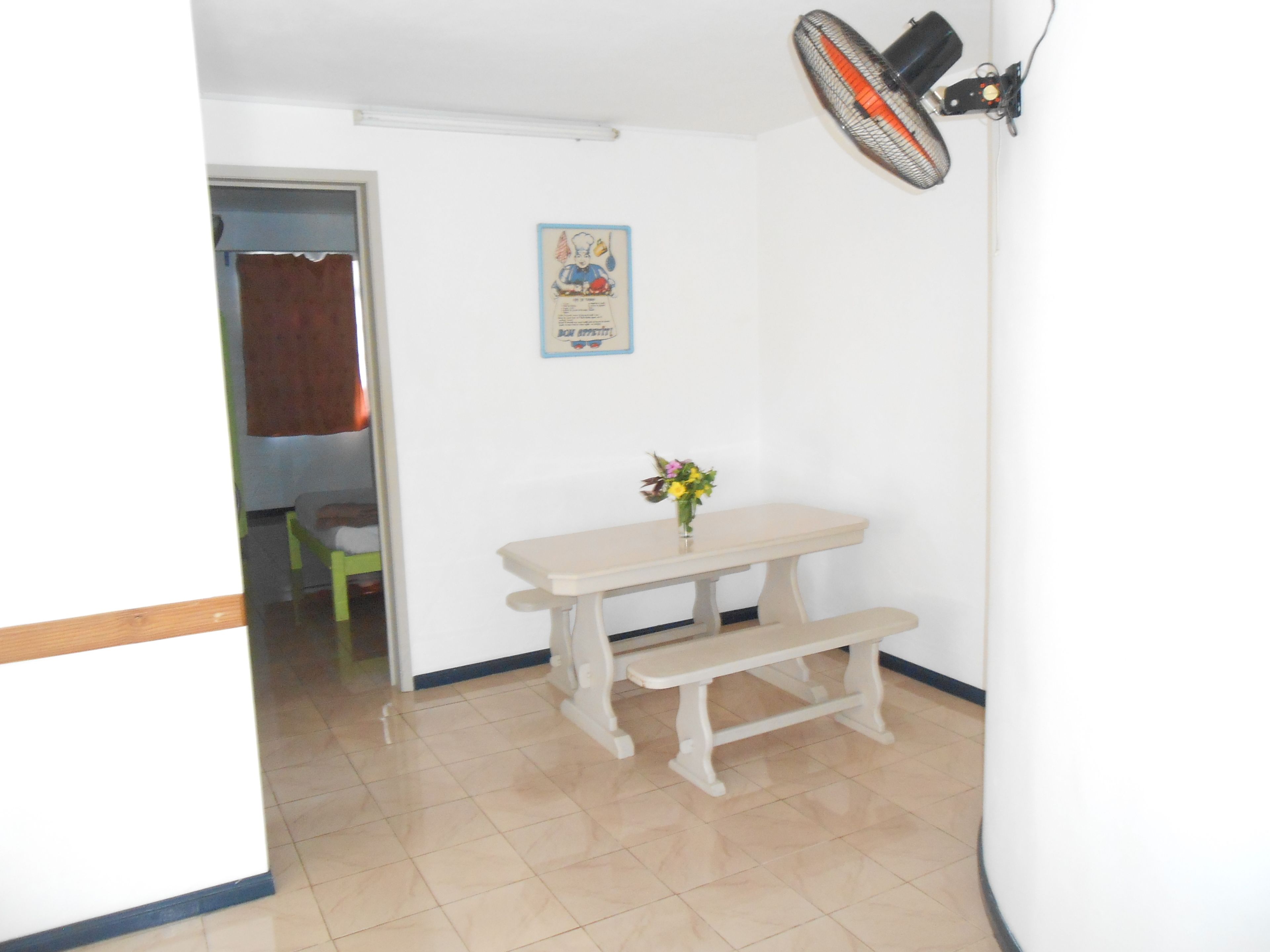 A small room with a white table and bench, a poster on the wall, and a fan mounted on the wall.